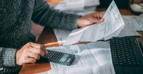 Een man achter een bureau met facturen op zijn bureau en een factuur in zijn hand maakt hij een berekening op zijn rekenmachine